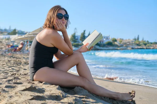Mujer de verano sentada en un libro de lectura en la orilla de arena, tomando el sol al atardecer — Foto de Stock