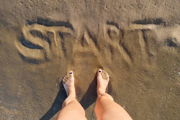 Texto de inicio escrito en arena en la playa del mar y las mujeres pies desnudos — Foto de Stock