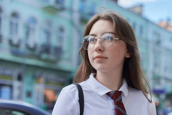Outdoor portrait of university student girl, confident female posing in white T-shirt, glasses, tie, backpack