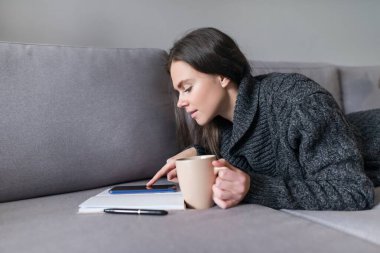 Genç bir kadın elinde aletlerle kanepede oturuyor dijital tabletli akıllı telefonlu akıllı telefon okuyan bir kadın bloglar, videolar, sosyal ağlar, fotokopi makinesi.