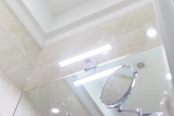 Bathroom interior, closeup detail. Part of the molding ceiling, LED light above the washbasin mirror, cosmetic makeup mirror, beige marble tiles on the wall