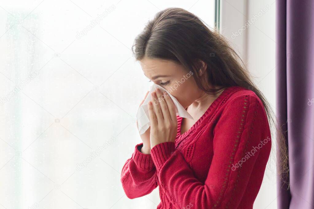 Ill young woman with handkerchief. Female with cough, snot, temperature standing at home near the window, copy space