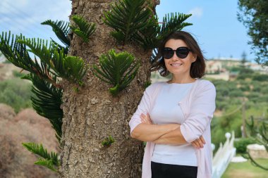 Kollarını kavuşturmuş orta yaşlı güzel bir kadın, tropik ağaçların yanında güneş gözlüklü bir kadın, fotokopi odası. Başarılı bir kadın dinleniyor.