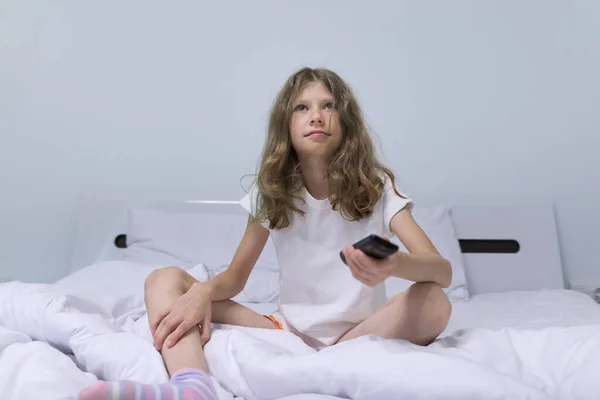早上漂亮的金发小女孩坐在白色的床上 带着电视遥控器 — 图库照片