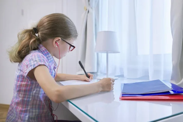 9歳の少女は 窓の近くの机に座ってノートを書き 耳のヘッドフォン オーディオレッスン 教育の技術 — ストック写真