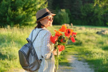 Yaz tatili, doğa gezisi, bir buket gelincik çiçeğiyle kır yolunda yürüyen olgun, güzel bir kadın manzaralı günbatımı arka planı, fotokopi alanı.