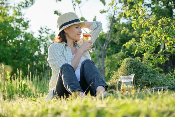 戴着帽子的女园丁坐在刚刚修剪好的草地上 用自制的天然饮料薄荷糖和草莓 绿色的花园背景 — 图库照片