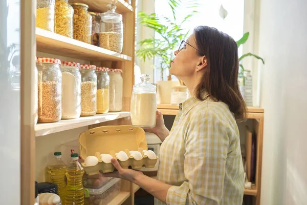 Woman in pantry taking products, eggs. Food storage, cooking at home, wooden shelf with grain products in storage jars