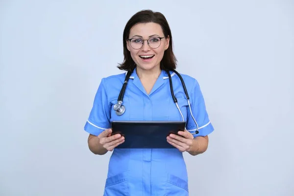 拥有数码平板电脑的成熟女性医务工作者 带着微笑的护士看着灰色背景的相机 — 图库照片