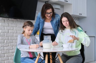 Anneleri mutfakta yemek yiyen çocuklar, iki kız ellerinde mısır gevreği ve sütle masada oturuyorlar. Aile, yemek, iletişim, sağlık konsepti.