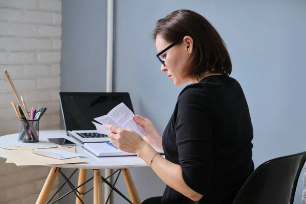 Mature business woman sitting at desk in office reading paper document, business correspondence, envelopes on table