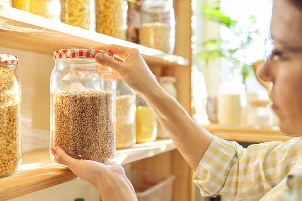 Food storage in pantry, woman holding jar of buckwheat in hand. Pantry interior, wooden shelf with food cans and kitchen utensils