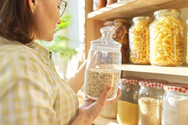 Food storage in pantry, woman holding jar of oatmeal in hand. Pantry interior, wooden shelf with food cans and kitchen utensils