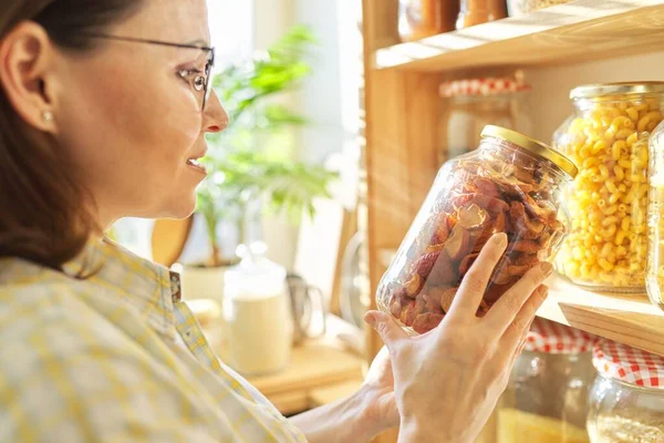 Food storage in pantry, woman holding jar of dry sun-dried apples in hand. Pantry interior, wooden shelf with food cans and kitchen utensils