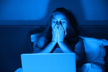 Genç bir kadın gece evde dizüstü bilgisayarla yatağında oturuyor. Kız başını elinde tutuyor, duygusal stres, kötü haber, depresyon, baş ağrısı