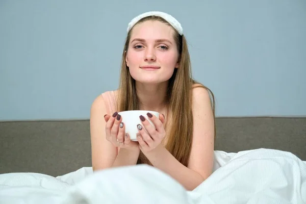 Beautiful young woman 20 years old blonde in bed with cup of tea, morning, white bed, background gray wall
