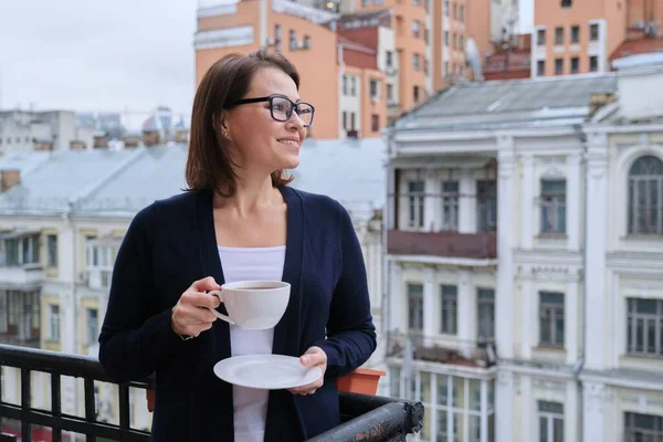Portrait of mature woman in glasses, cardigan with cup of tea on open balcony in city. Background urban architecture, buildings, smiling female looks away, copy space