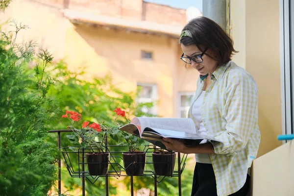 Beautiful mature woman in home clothes on open balcony reading magazine — Stock Photo, Image