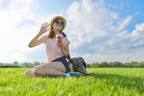 ファッショナブルな夏の肖像画の十代の女の子の帽子とともにドリンク座って緑の草 — ストック写真