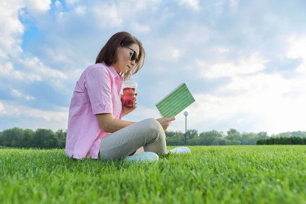 Femme mûre lisant le livre, femme assise sur l'herbe verte avec boisson — Photo
