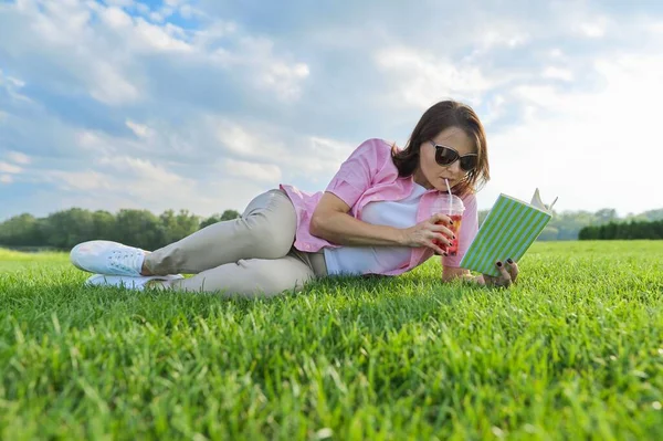 Dojrzała kobieta czyta książkę, kobieta leży na zielonej trawie z napojem — Zdjęcie stockowe