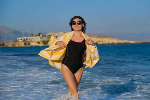 Щаслива зріла жінка в купальнику з пляжним рушником, що йде уздовж моря — стокове фото
