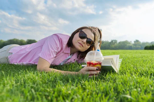 Mature woman reading book, female lying on green grass with drink