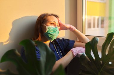 Tıbbi yüz maskesi takan doktor kadının portresi, kliniğin penceresine bakıyor.