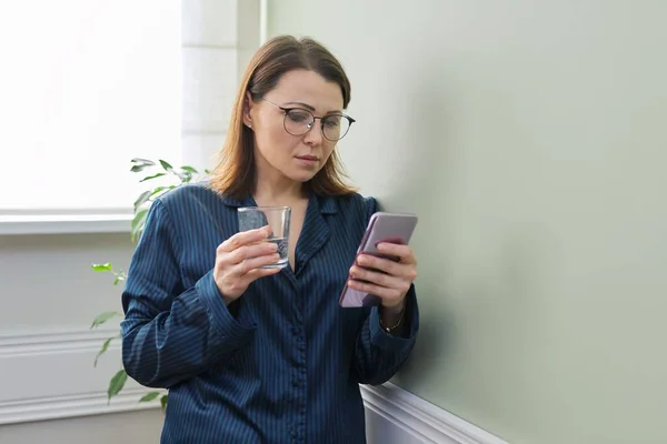 Morgonporträtt av mogen vacker kvinna med glas vatten och smartphone — Stockfoto