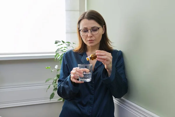 Reife Frau tropft Tropfen mit Glas Wasser, trinkt Beruhigungsmittel, zu Hause im Schlafanzug — Stockfoto