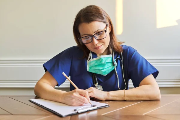 Жінка-лікар у блакитній формі, медична маска сидить за столом, пишучи на кишені — стокове фото