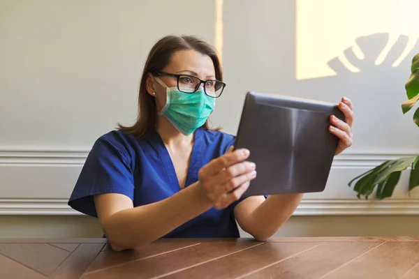 Retrato de doctora en máscara de protección médica con tableta digital sentada en el escritorio en la oficina — Foto de Stock