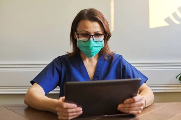 Retrato de doctora en máscara de protección médica con tableta digital sentada en el escritorio en la oficina —  Fotos de Stock
