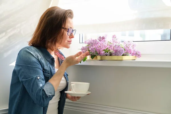 Mujer de pie cerca de la ventana con taza de té y ramo de lilas, espacio para copiar —  Fotos de Stock