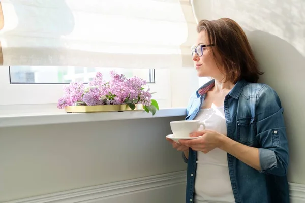 Mujer de pie cerca de la ventana con taza de té y ramo de lilas, espacio para copiar — Foto de Stock