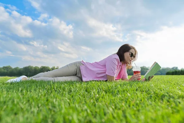 Femme mûre lisant le livre, femelle couchée sur l'herbe verte avec boisson — Photo