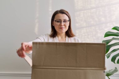 Mature woman unpacks cardboard box on table at home, in office clipart