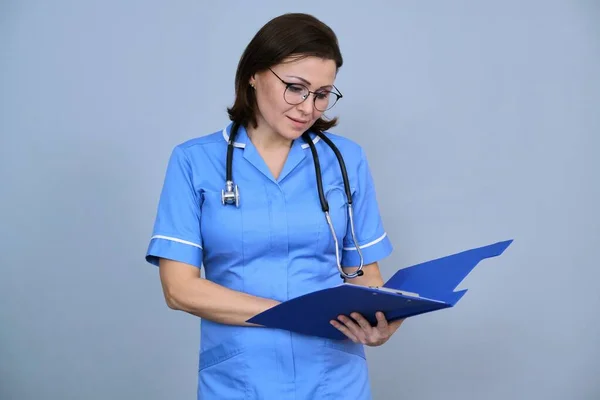 成熟护士拿着剪贴板的画像 身穿蓝色制服的女性 听诊器检查灰色背景的剪贴板 — 图库照片