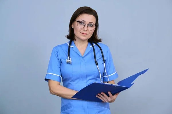 成熟护士拿着剪贴板的画像 身穿蓝色制服的女性 带听诊器 看着灰色背景的相机 — 图库照片