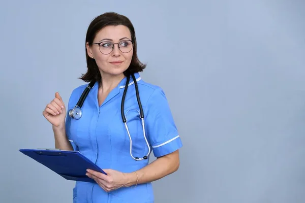 成熟护士拿着剪贴板的画像 蓝色制服的女性 灰色背景听诊器 复制空间 — 图库照片