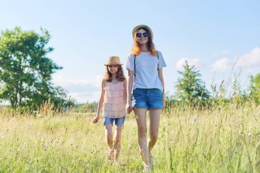 Yaz tatilleri, çocuklar, iki kız kardeş el ele yürüyorlar, mavi gökyüzü çayırları. Aktif sağlıklı yaşam tarzı, dost canlısı bir aile.