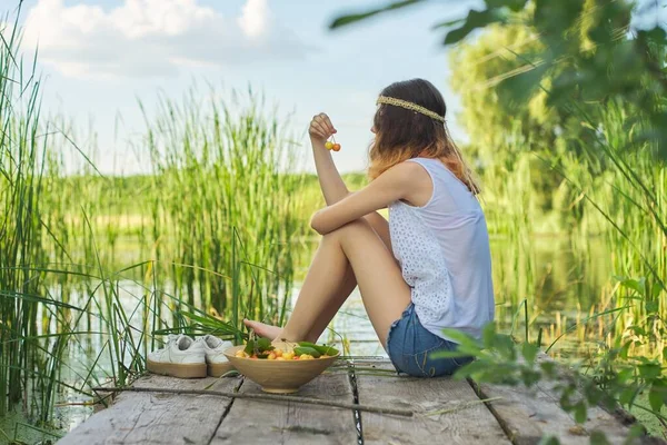 少女坐在木制码头上 戴着黄色的樱桃 美丽的年轻女子在湖上吃着天然的水果 欣赏着美丽的落日 健康食品 复制空间 — 图库照片