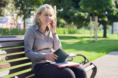 Kendine güvenen olgun bir kadın cep telefonuyla konuşuyor, şehirde başarılı bir kadın bankta oturuyor, iş not defterine bakıyor.