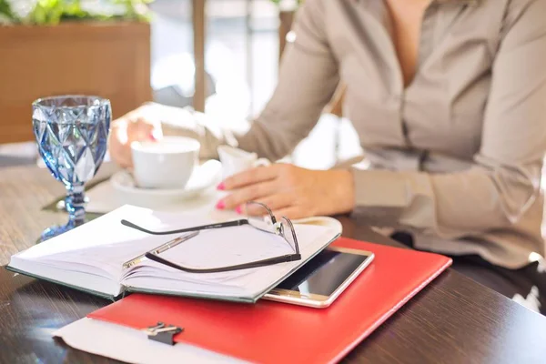 Coffee break. Table in an outdoor restaurant, business papers, notebook, smartphone, glasses, cup of coffee. Businesswoman in defocus