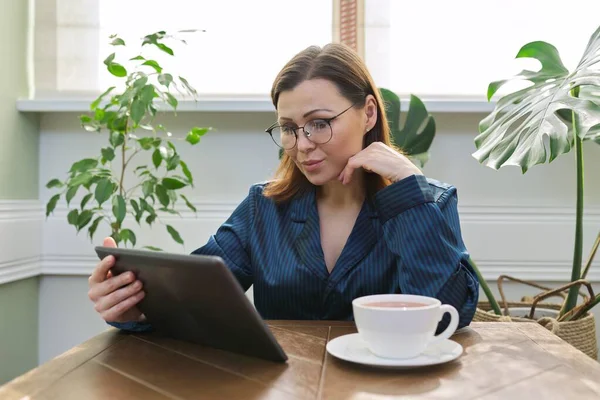 自宅読書メール ニュース デジタルタブレット上のソーシャルネットワークで成熟した深刻な女性 朝の女性のパジャマでお茶を飲んで — ストック写真