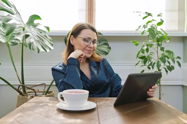 テーブルでデジタルタブレットを読んで座ってお茶のカップとパジャマで自宅で朝食を持って笑顔成熟した女性 メールを読んでくれた幸せな女性ニュース 肯定的な感情 40人 — ストック写真