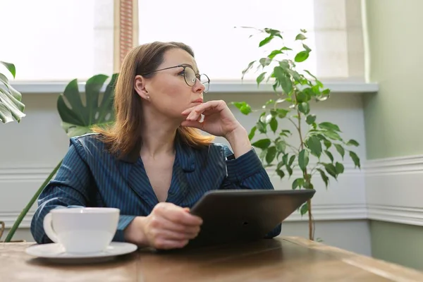 深刻な成熟した女性は テーブルのデジタルタブレットを読んで座ってお茶を飲みながらパジャマで自宅で朝食を持っています 悲しい女性はメールを読んで ニュース 否定的な感情 中年の人々の生活 — ストック写真