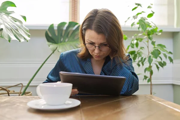 深刻な成熟した女性は テーブルのデジタルタブレットを読んで座ってお茶を飲みながらパジャマで自宅で朝食を持っています 悲しい女性はメールを読んで ニュース 否定的な感情 中年の人々の生活 — ストック写真