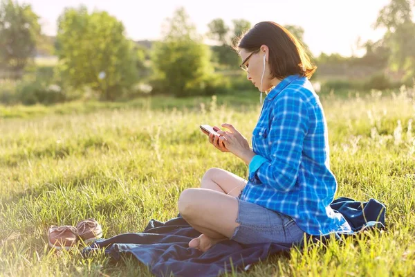 成熟的女人听音乐 耳机上的有声读物 在自然界中放松 背景日落 乡村风景 绿色草地 复制空间 — 图库照片