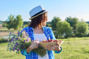Bir kase taze toplanmış çilek ve bir buket kır çiçeğiyle kadın çiftçi. Doğa arka planı, kırsal alan, yeşil çayır, kır tarzı.